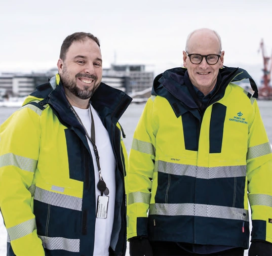 Two peaople with yellow jackets straning in the port area looking into the camera.